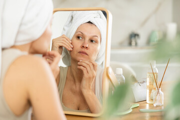 Attentive woman in her middle age inspecting her face in a mirror sitting at a table in home environment