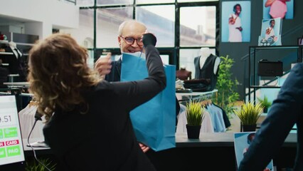 Retail assistants selling clothes to elderly client at fashion boutique, giving discount coupon to regular customer. Employees team assisting senior man to buy clothing items on sale. Camera B.
