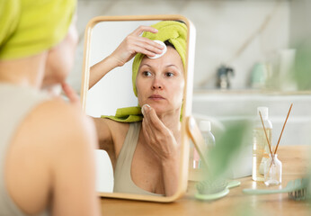 Focused and calm woman with towel wrapped around head attentively removing makeup with cotton pads...