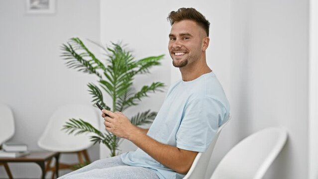 A smiling young hispanic man with a beard in a blue shirt sits holding a phone in a bright waiting room.