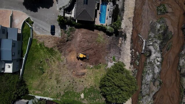 Drone shot of a yellow digger clearing a site in preparation for building next to a river, other houses and a large tree