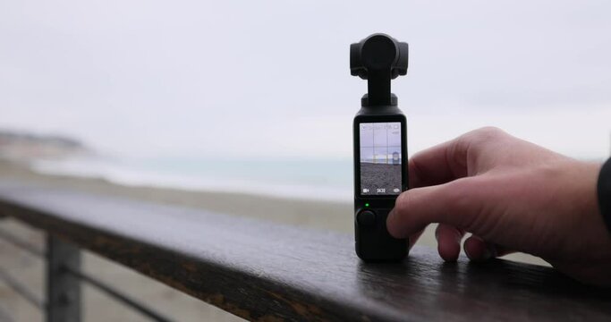 Portable 3-Axis Stabilized 4K Handheld Camera Being Turned On As It Rests On Bannister Beside Beach With Bokeh Background. Static Shot