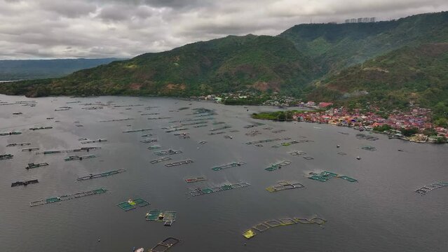 Crowded Tilapia Fish Cages on Taal Lake in Philippines. Mountain landscape and small village at coast. Cloudy day in Asia. Aerial forward flight.