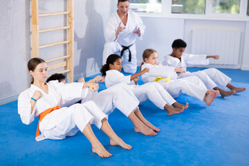Multinational group of focused teenagers in white kimono performing core twist exercise with...