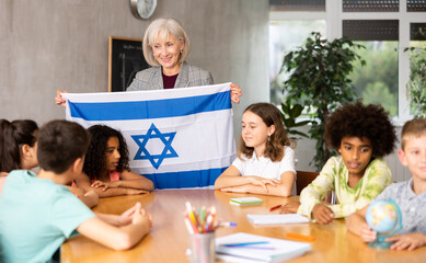 Mature woman teacher tells pupils the history of Israel in class and holds the national flag of the...