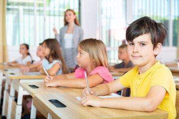 Portrait of smiling tween schoolboy sitting in classroom during lesson, writing exercises in workbook ..