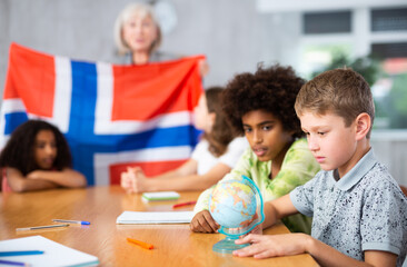 Woman teacher working in a high school tells pupils about Norway in a history lesson and holds the country national flag