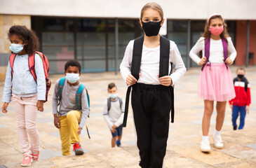 Portrait of positive schoolgirl in mask standing near school, children on background