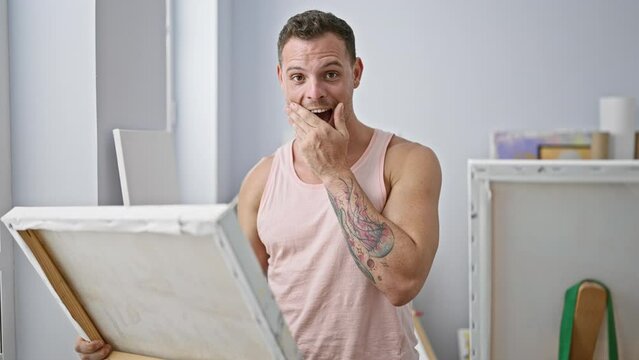 A surprised young man with tattoos in a tank top reacts emotionally while holding an empty canvas in a bright studio