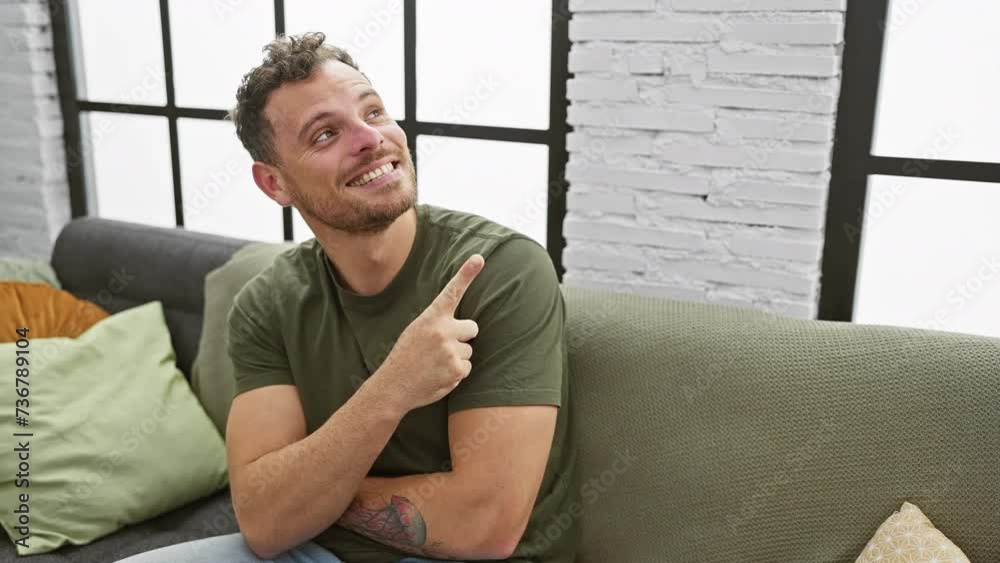 Sticker Smiling, optimistic young man, confidently pointing off to the side while sitting on his living room sofa at home, a joyous expression on his handsome face.