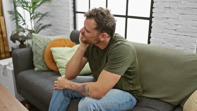 Depressed young man, bored and tired, reflectively sitting inside, arms crossed, lost in tedious thinking on sofa at home