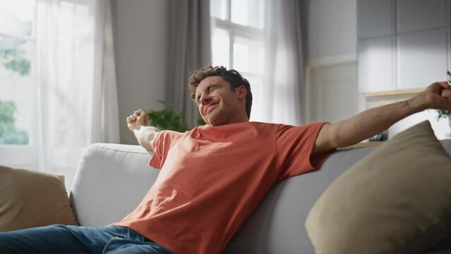 Chilling guy sitting sofa stretching arms at home. Relaxed man taking smartphone