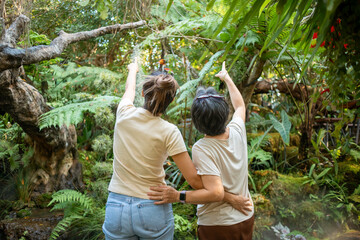 Back side of asian Thai daughter and elder mother standing and enjoying natural forest garden together, pointing at bird, traveling on holidays.