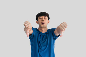 Fototapeta premium A young Asian man in his 20s wearing a blue t-shirt making a displeased gesture showing a thumbs down isolated on a gray background. The concept of expressing dislike or opposing opinions.