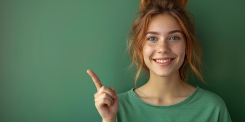 A smiling young woman with freckles pointing up, standing against a green background, evoking a sense of cheerfulness and positivity.