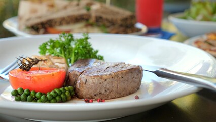 Grilled steak and vegetables on white plate, elegantly presented with side of greens and tomato, served at outdoor restaurant setting. Gourmet dining and presentation.