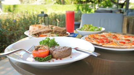Gourmet outdoor dining experience with variety of dishes including grilled steak, garden salad, sandwiches, and pizza. Outdoor restaurant eating and culinary diversity.