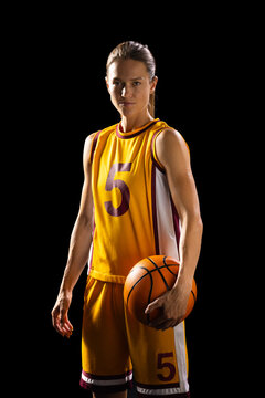 Athletic young Caucasian female basketball player poses confidently in basketball gear on a black ba