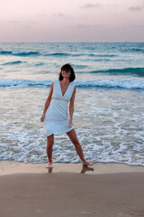 Fototapeta na wymiar Happy woman in a white dress enjoys relaxing on the beach, walking along the sandy shore at sunset.