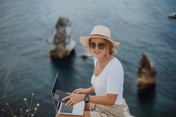 Freelance women sea working on the computer. Good looking middle aged woman typing on a laptop keyboard outdoors with a beautiful sea view. The concept of remote work.