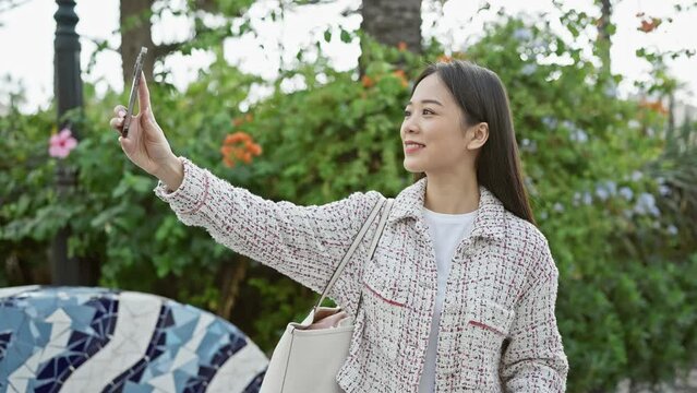 A young asian woman in a casual tweed jacket taking a selfie with her smartphone in a verdant urban park.