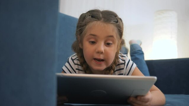 Little kid girl using tablet device, lying on sofa. Smiling little girl watching online cartoons on tablet on Internet using digital tablet technology device. kid plays at home sofa computer addiction