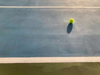 the green tennis ball is in super bright natural daylight showing its shadow on a blue-green tennis court