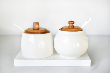 A salt shaker and a sugar bowl in white with wooden lids stand on a white table. Elements of decor in a modern interior. Modern White Tableware