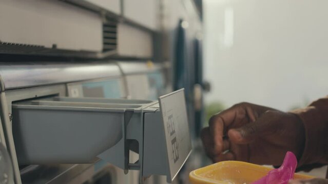 Side close-up of hand of anonymous man adding one spoon of powder detergent into washing machine drawer before starting laundering at washateria