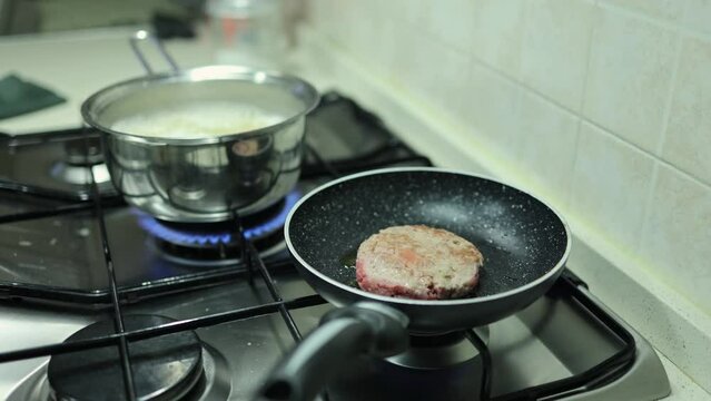 Burger Cooked in a Pan on a Home Kitchen Stove and a Pot with Pasta That Boils