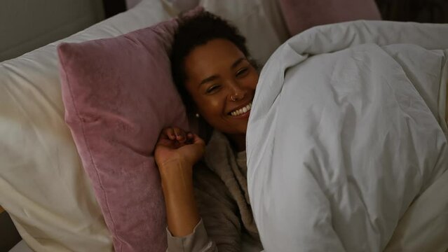 A smiling woman with curly hair comfortably nestled in bed, surrounded by pink pillows and white sheets, in a cozy bedroom setting.