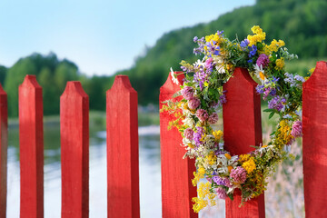 colorful flowers wreath on red wooden fence outdoor. summer background. Floral crown, symbol of Summer Solstice Day, Midsummer holiday, Litha sabbath. witch, wiccan ritual. copy space