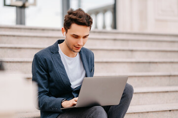 Concentrated young man wearing ear-pods is working on laptop while sitting outdoors.