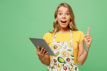 Young smart housewife housekeeper chef cook baker woman in apron yellow t-shirt use digital tablet pc computer look for recipe point finger up isolated on plain green background Cooking food concept