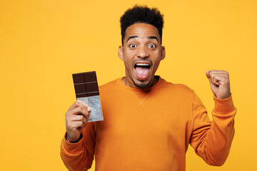 Young surprised man wear orange sweatshirt casual clothes hold eat bar of chocolate do winner...