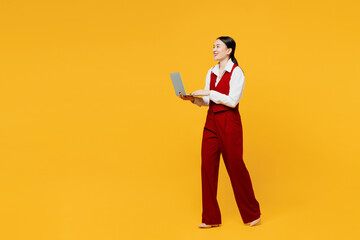 Full body side view smart young lawyer employee IT business woman of Asian ethnicity in formal red vest shirt work at office go hold use laptop pc computer isolated on plain yellow background studio