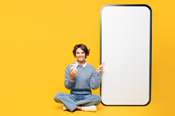 Full body young woman wears grey knitted sweater shirt casual clothes sit point on big huge blank screen area mobile cell phone use smartphone isolated on plain yellow background. Lifestyle concept.