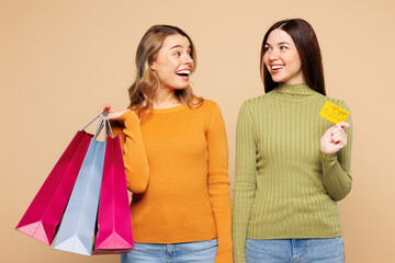 Young fun friends two women wear orange green shirt casual clothes hold in hand credit bank card shopping open paper package bags isolated on plain beige background. Black Friday sale buy day concept.