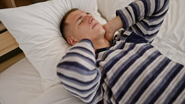 A young man in a striped shirt appears uncomfortable as he clutches his neck lying in bed, indicating pain or soreness, indoors at home.