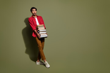 Full length photo of shocked funky guy wear red shirt walking holding heavy book stack empty space...