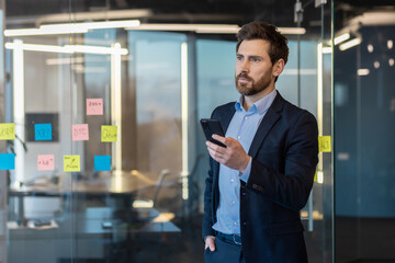 Professional mature businessman in a modern office, intently using a smartphone with strategy notes visible on glass.