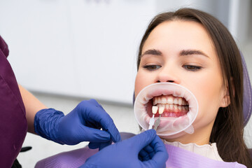 The dentist meticulously chooses the enamel shade for the teeth of the young brunette woman before...
