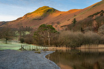 Winter morning sunlight on Catbells mountain peak seen from the shoreline of Derwentwater in The Lake District, UK.