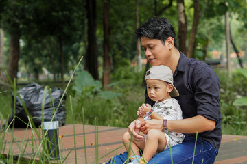 Father and son sitting at the park, father carrying son, surrounded by green grass.