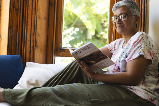 A mature biracial senior woman enjoys reading a book at home, with copy space
