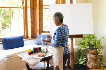 A mature biracial woman paints on a canvas at home, with copy space