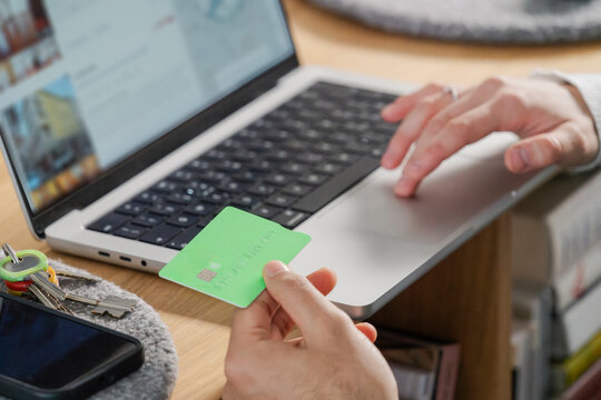Close-up of a man's hand holding a green credit card over a laptop, finalizing an online payment
