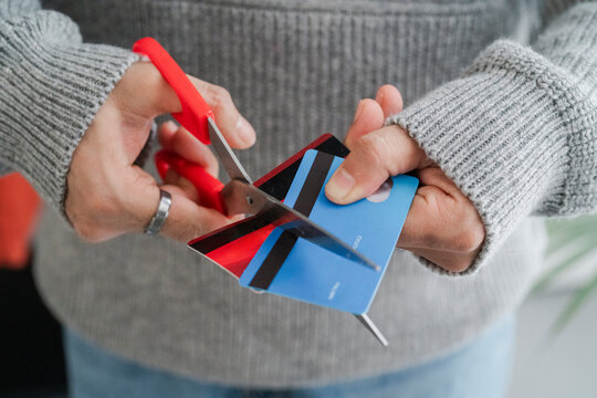 A man's hands cutting a blue credit card with red scissors, symbolizing financial management or the end of a credit account. The decisive cut reflects personal finance decisions