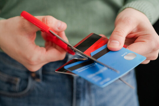 A person in a green sweater is seen cutting a stripe-patterned credit card with red scissors. The act signifies the intention to reduce debt or prevent overspending