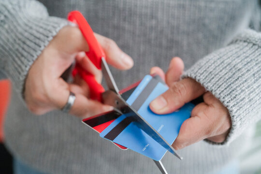 A man's hands cutting a blue credit card with red scissors, symbolizing financial management or the end of a credit account. The decisive cut reflects personal finance decisions